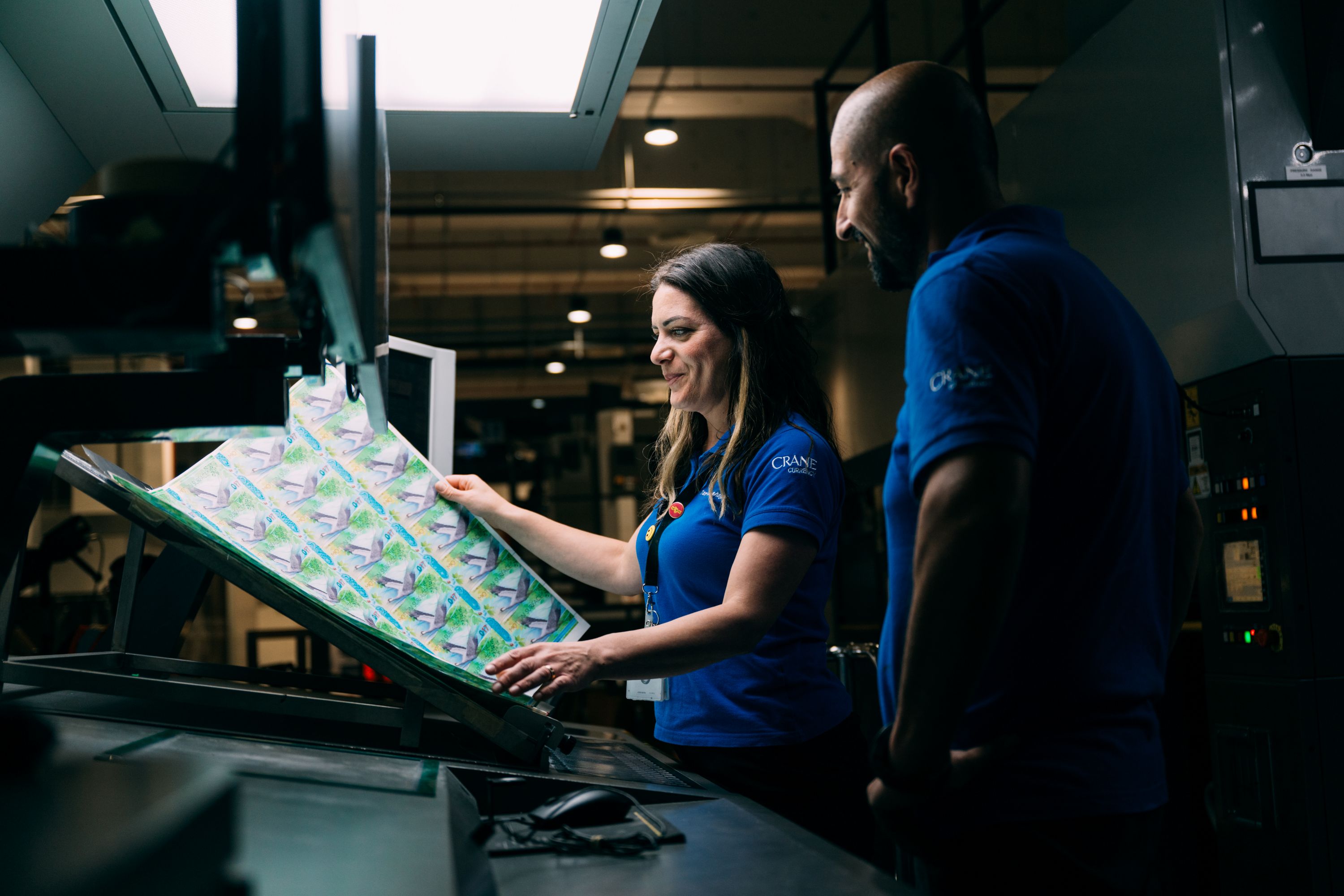 A photo of "the Beauty of Life" house note in production with two Crane Currency associates inspecting a banknote sheet.
